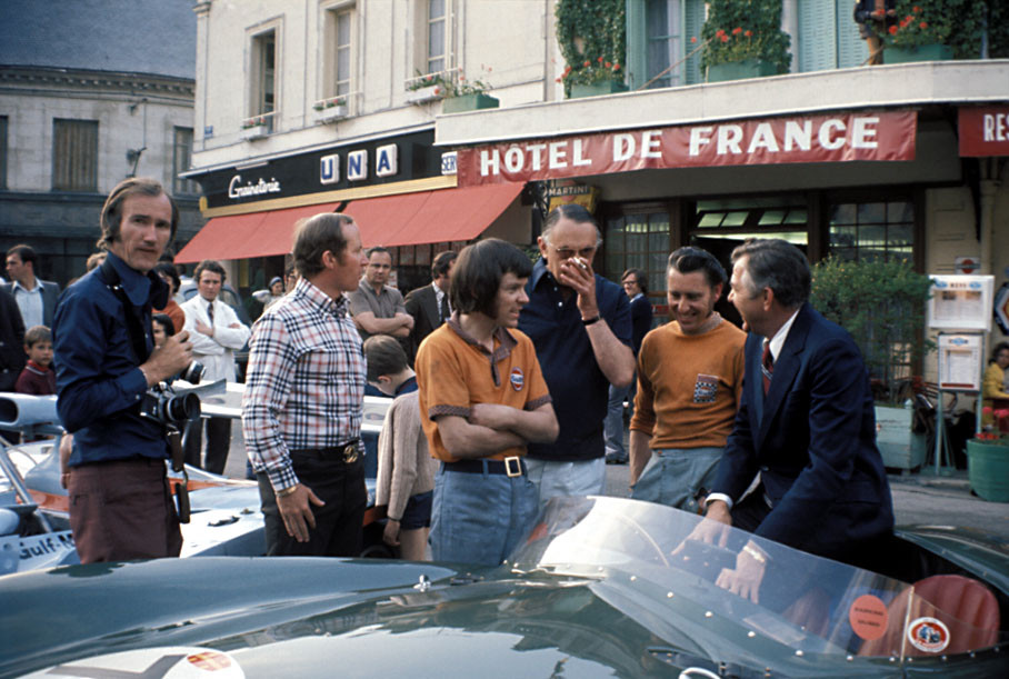 Photographer Nigel Snowdon approaches the group of John Wyer and Gulf mechanics Robin and Alan next to the Gulf Mirage Ford and an Aston Martin DB3 with its owner Richard Pilkington in front of Hôtel de France in Chateau du Loir